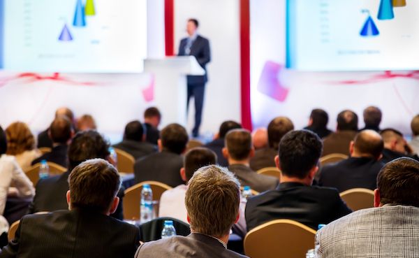A speaker on stage addressing a full conference audience.